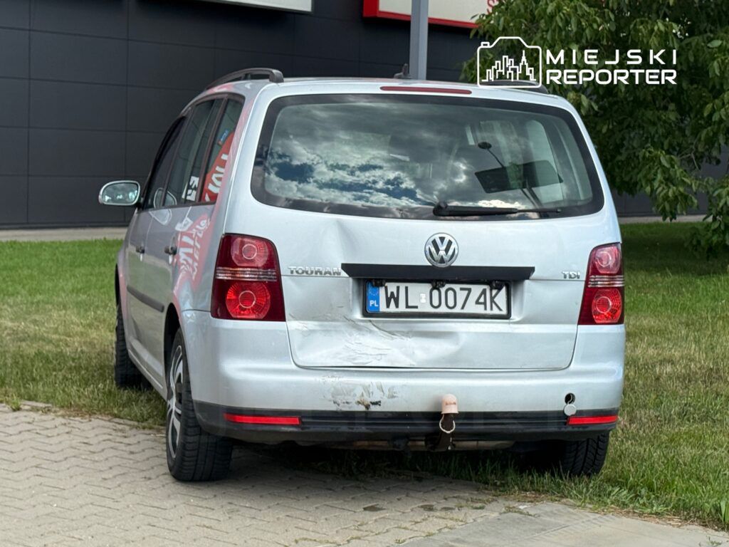 Uszkodzony srebrny Volkswagen Touran zaparkowany na trawie obok nowoczesnego budynku.