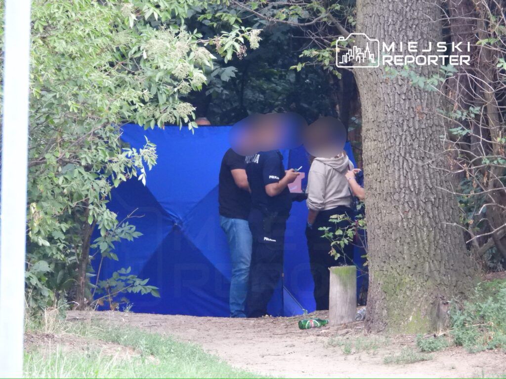 Grupa policjantów rozmawia z osobą w szarej bluzie przy niebieskim namiocie w leśnym otoczeniu.