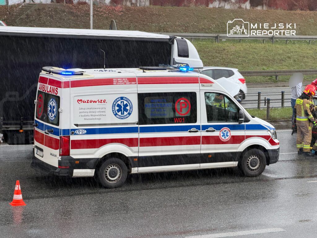Karetka pogotowia z włączonymi sygnałami świetlnymi stoi na drodze, obok której pracują strażacy.