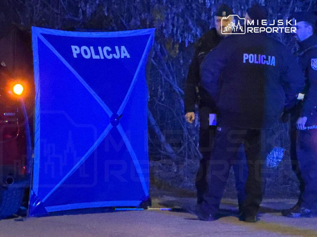 Grupa policjantów w mundurach rozmawia obok niebieskiego namiotu z napisem "POLICJA" na ciemnej ulicy.