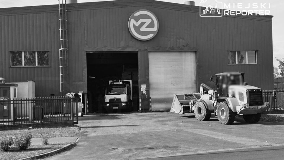 Białe ciężarówki i koparka zaparkowane przed dużym, szarym budynkiem przemysłowym z logo na fasadzie.