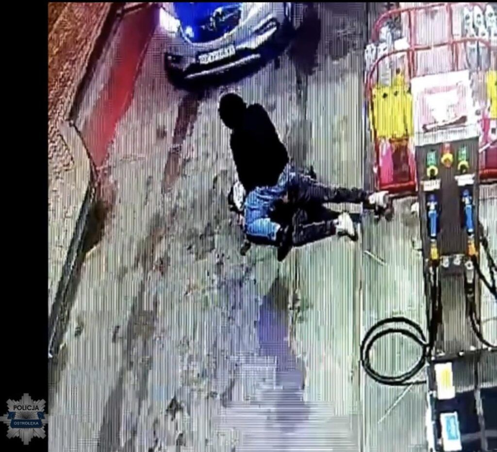 Mężczyzna w czarnej bluzie przemocą obezwładnia innego mężczyznę na stacji paliw, obok stoją samochody.