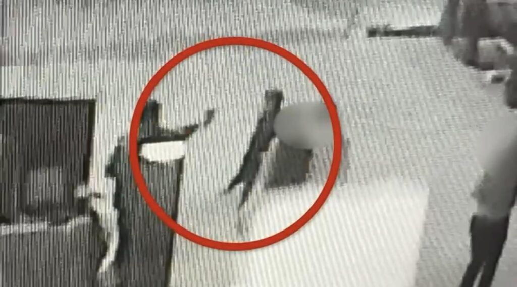 Osoba w ciemnym ubraniu trzyma przedmiot w ręku, stojąc w zamkniętym pomieszczeniu z widocznymi innymi osobami.