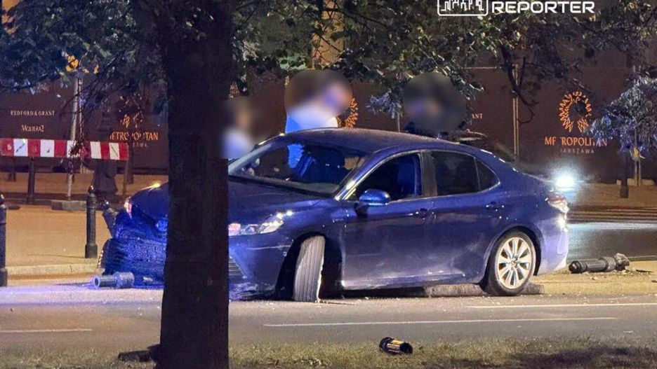 Niebieski samochód osobowy z uszkodzoną oponą zaparkowany na ulicy, obok leżące przedmioty. W tle widoczne drzewa i oświetlenie nocne.