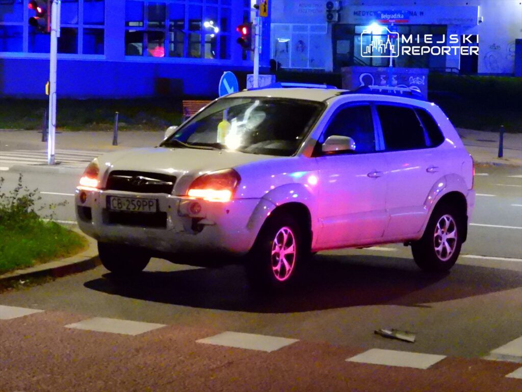 Biały SUV zatrzymany na skrzyżowaniu, z włączonymi światłami, w nocy, w pobliżu budynku z dużymi oknami.