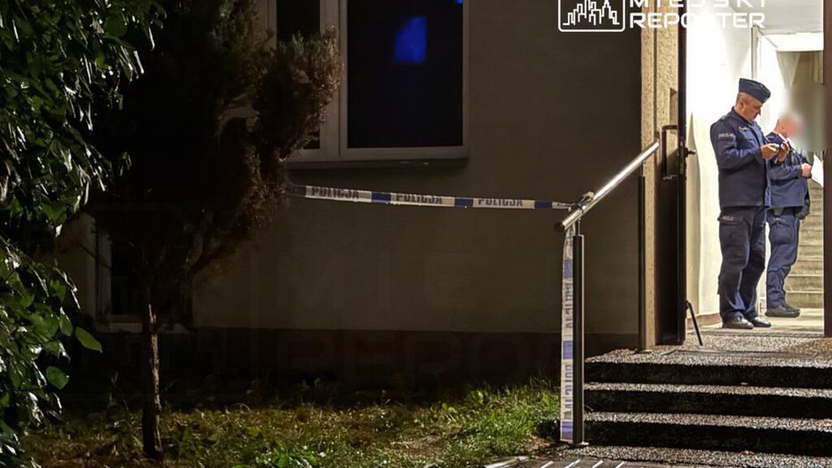 Dwóch policjantów stojących przy wejściu do budynku, obok taśmy zabezpieczającej. Nocna sceneria z roślinnością w tle.