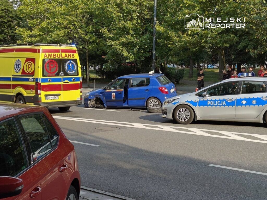 Ambulans i radiowóz policyjny stoją obok uszkodzonego niebieskiego samochodu na ulicy, w tle grupka ludzi.