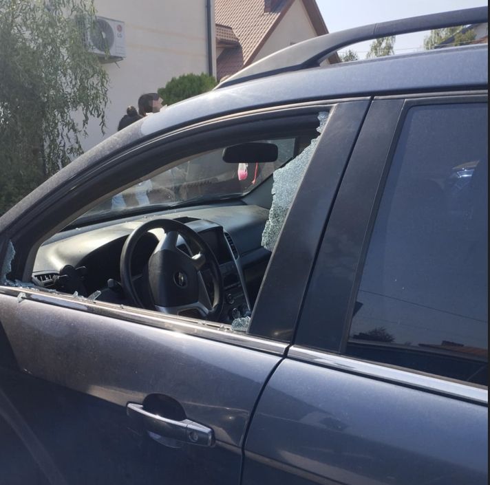 Uszkodzone auto z wybitym oknem, w środku widoczna kierownica, w tle osoby stojące obok budynku.
