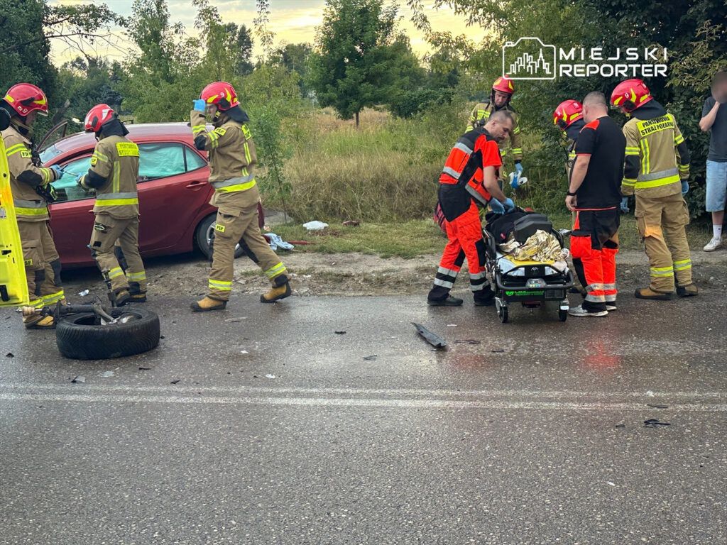 Sześciu strażaków w hełmach i mundurach udziela pomocy przy rozbitej czerwonej osobówce, obok leży odcięte koło.