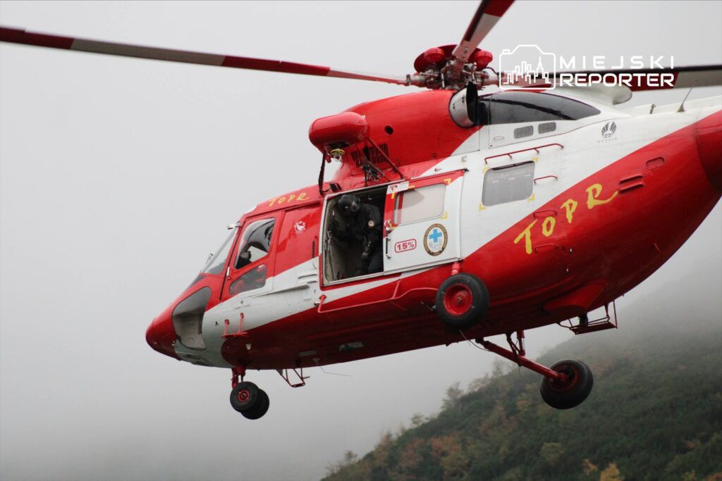 Helikopter ratunkowy w czerwono-białych barwach w trakcie lotu nad górskim terenem, z otwartymi drzwiami.