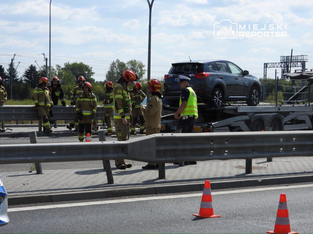 Grupa strażaków w uniformach omawia sytuację na poboczu drogi, obok uszkodzonego samochodu na lawecie.