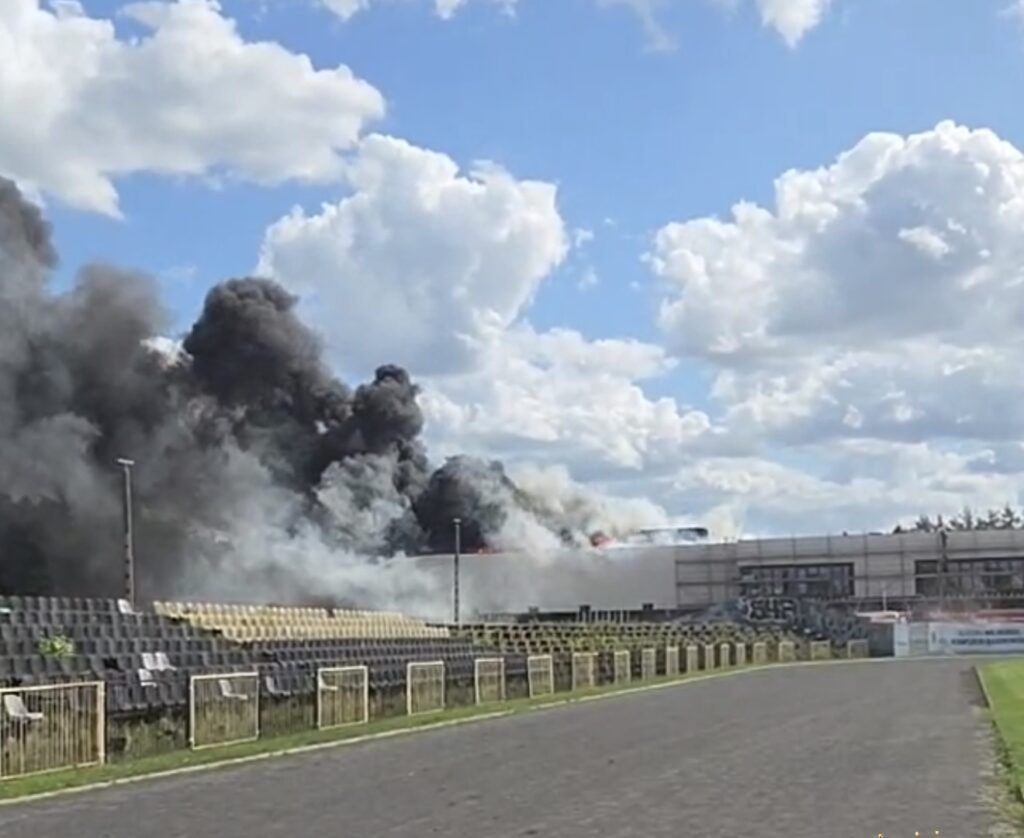 Gęsty czarny dym unoszący się nad stadionem, w tle widoczny budynek z płomieniami.