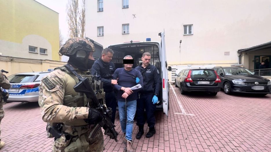 Mężczyzna w kajdankach prowadzony przez policjantów na parkingu, w tle radiowóz i inne samochody.