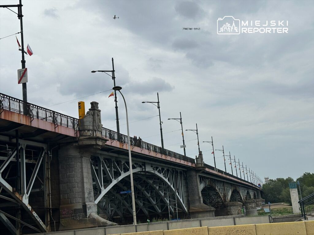 Most z metalową konstrukcją z widocznymi filarami, nad którym widać flagi, a w tle przelatujący samolot.