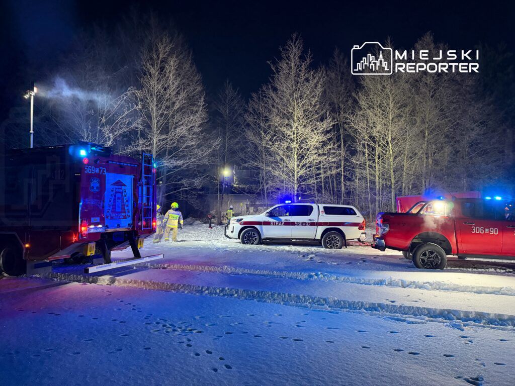 Strażacy w żółtych mundurach pracują nocą w zaśnieżonym lesie, obok pojazdów ratunkowych z włączonymi sygnałami świetlnymi.