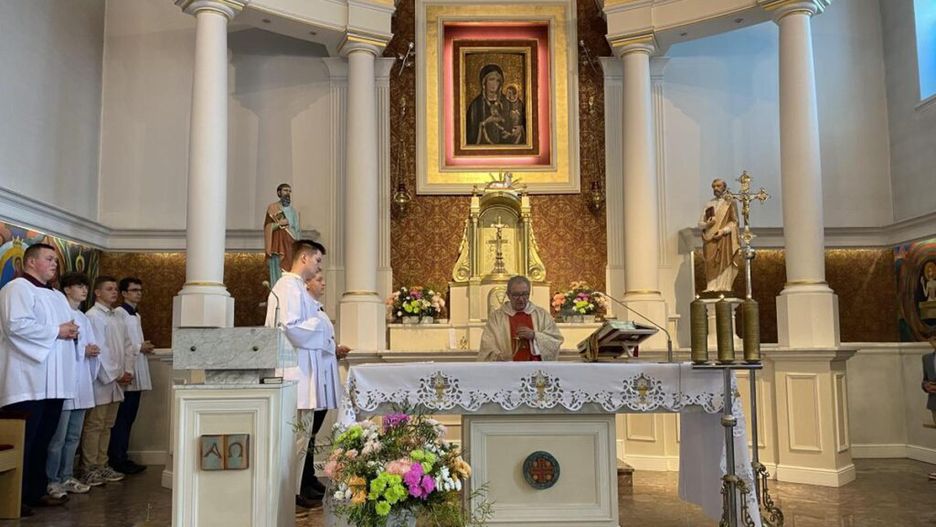 Ksiądz w szatach liturgicznych odprawia mszę przy ołtarzu w kościele, a wierni stoją w skupieniu.