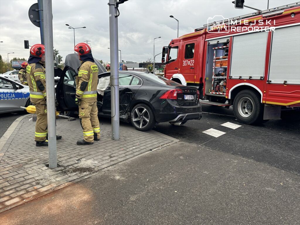 Dwaj strażacy w mundurach z hełmami rozmawiają obok uszkodzonego samochodu osobowego na skrzyżowaniu, w tle widać wóz strażacki.
