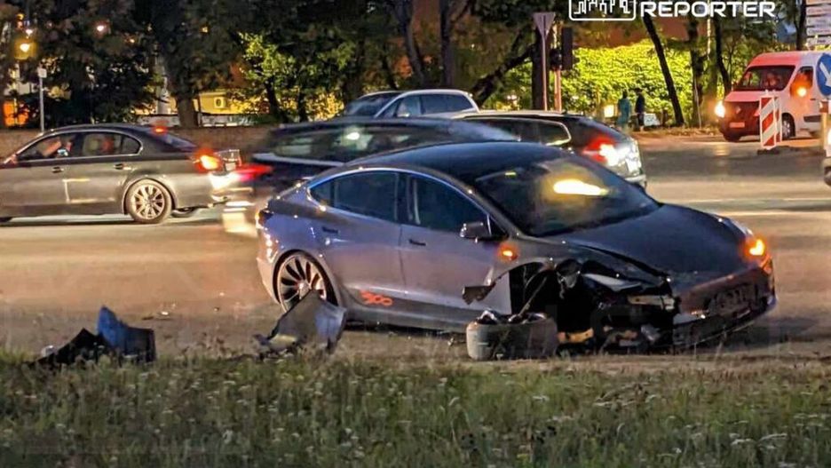 Zniszczony samochód osobowy stoi na poboczu drogi, obok przejeżdżające pojazdy w ruchu nocnym.