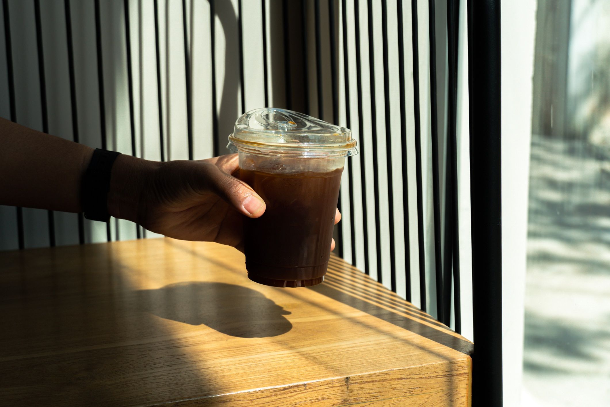 Hand holding black ice Coffee in drinking glass on wood table background with a sunbeam from the window. Summer refreshing drink. Black coffee in the morning with sunlight.
SandyHappy