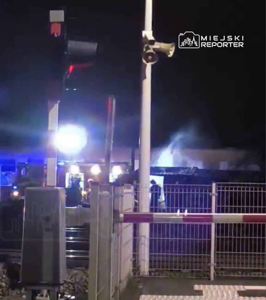 Na stacji kolejowej strażacy gaszą pożar, a w tle widać migające niebieskie i czerwone światła.
