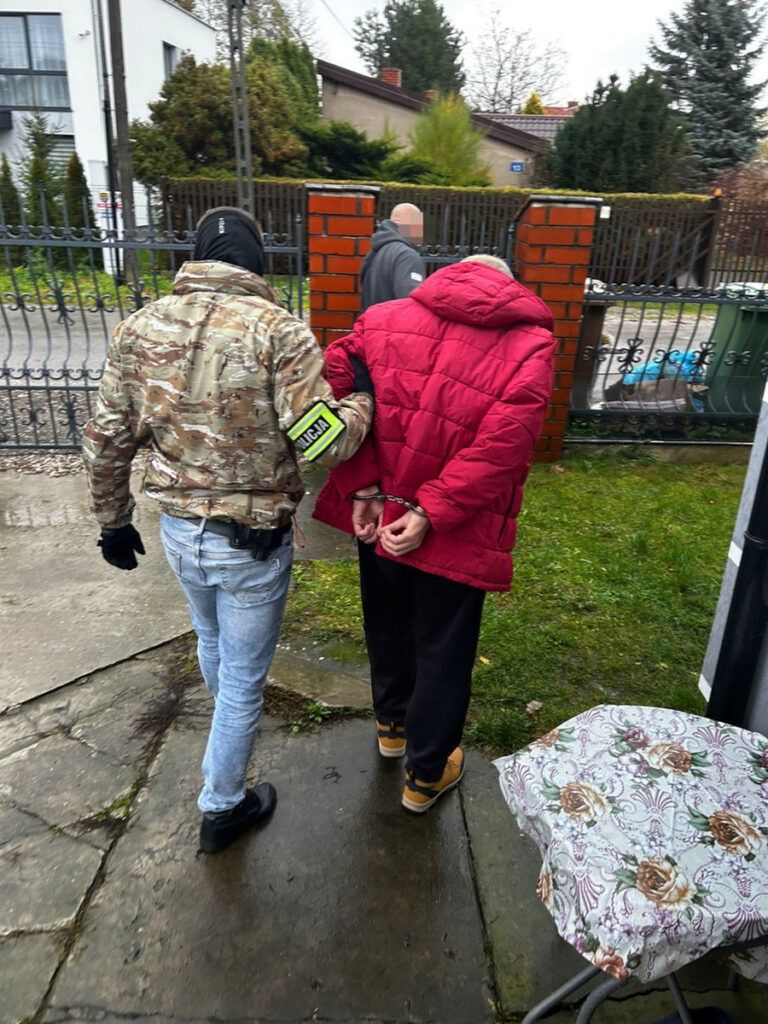 Funkcjonariusz w kamizelce taktycznej prowadzi aresztowanego mężczyznę w czerwonej kurtce przez posesję.