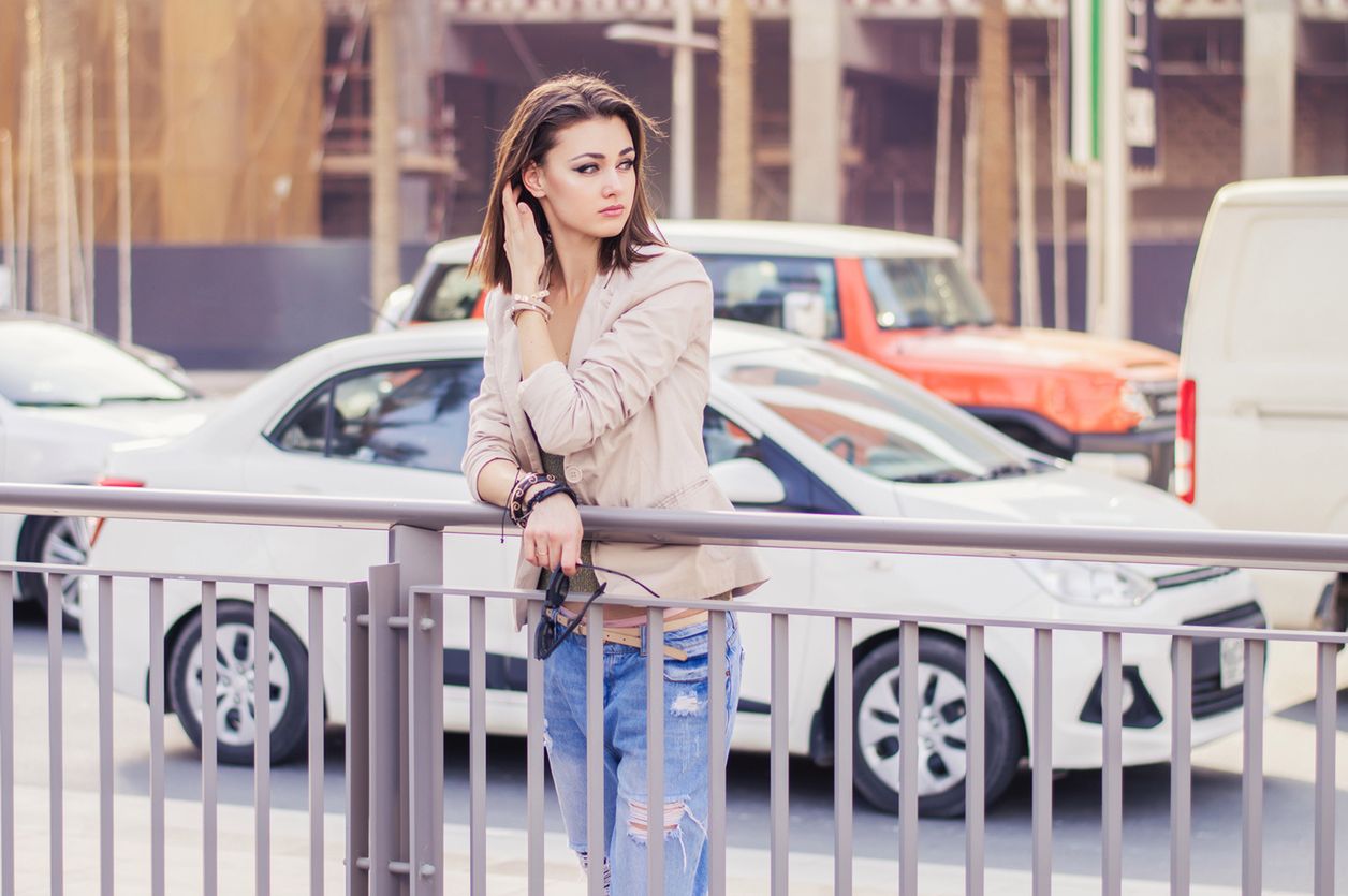 Young european female with trendy bob cut hairstyle standing near busy road in city center. Female in beige jacket and blue jeans standing in Dubai downtown 