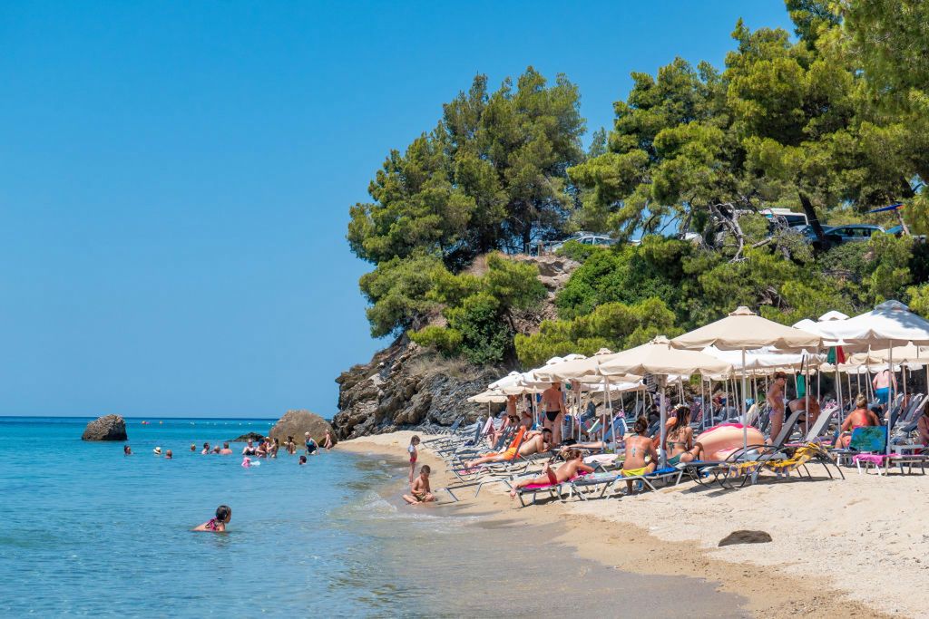 Sandy Beach and beach bar with sunbeds and umbrellas called Tortuga at Sithonia peninsula, Halkidiki, Greece on 17th July 2019. The beach with the crystal clear clean seawater under the summer sun with tourists and visitors from around the world swimming, sunbathing - tanning and relaxing in the turquoise Mediterranean water of Aegean Sea, as Chalkidiki attracts worldwide tourism because of the hotel infrastructure and nature. The beach is in a beautiful natural point below the shadow of Pine trees forest and Kelifos island is visible. The location is between Lagomandra and Tripotamos near Nikiti and Neos Marmaras. (Photo by Nicolas Economou/NurPhoto)