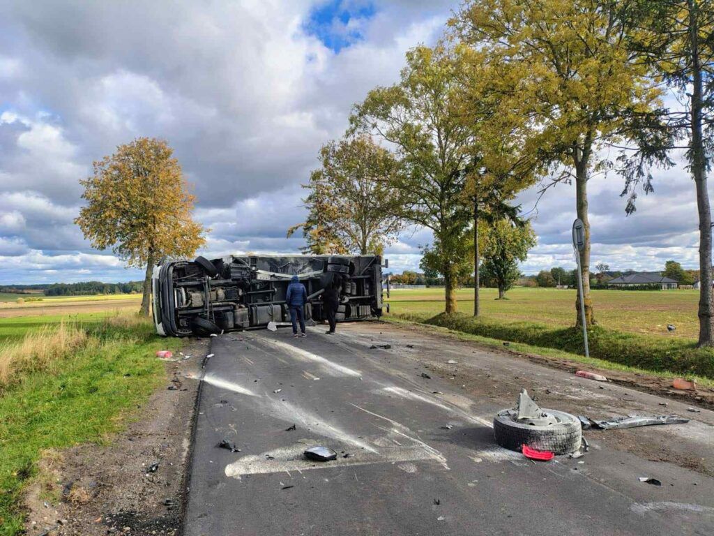 Przewrócony pojazd ciężarowy na drodze, obok dwaj mężczyźni zbierający pozostałości w otoczeniu pól i drzew.