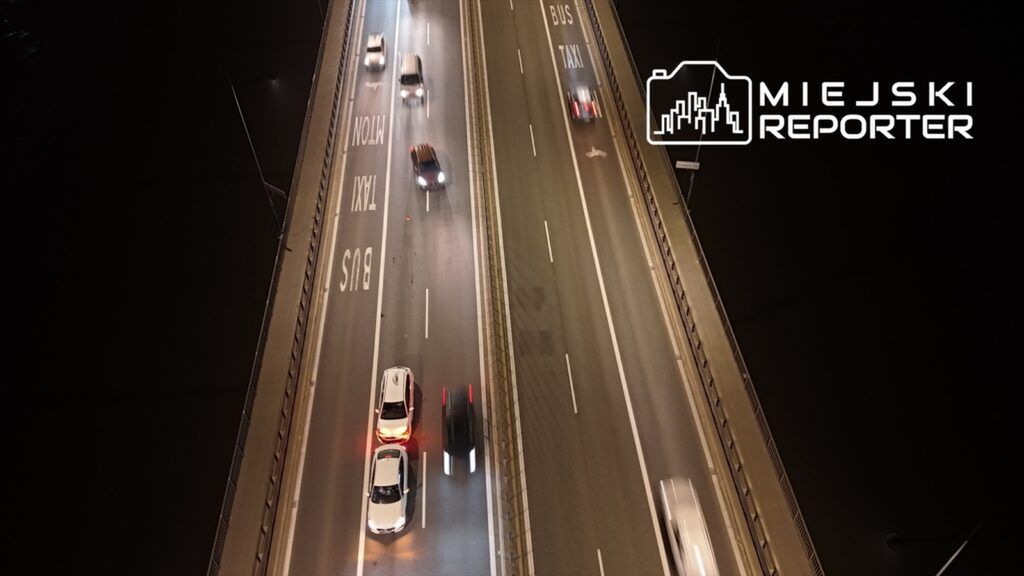 Ruch samochodów na oświetlonym moście z oznaczeniami drogowymi dla busów i taksówek.