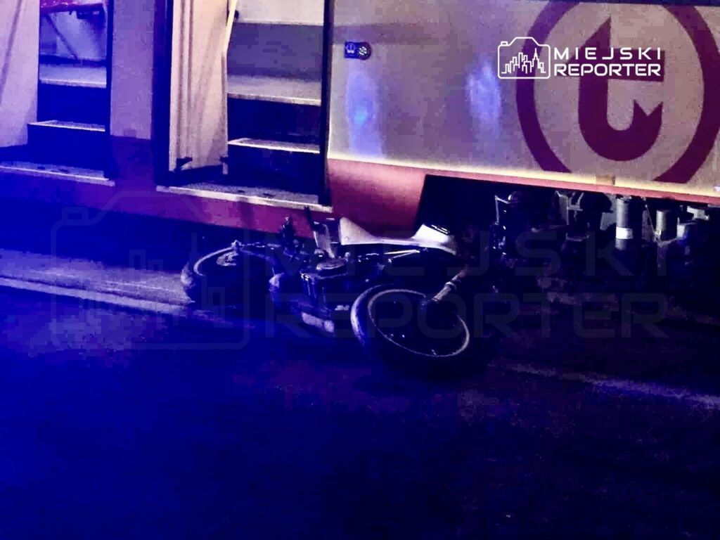 Motocykl leży na drodze obok tramwaju, w tle widać niebieskie światła policyjne.