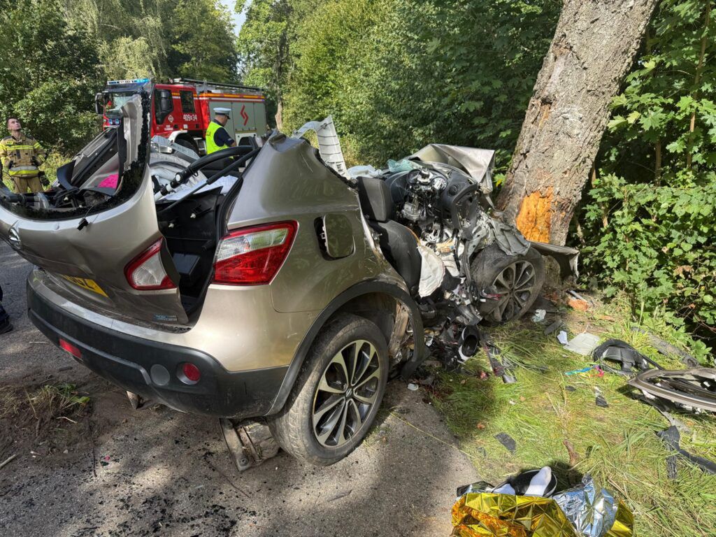 Rozbity samochód osobowy w kolorze złotym stoi przy drzewie w lesie, otoczony przez służby ratunkowe.
