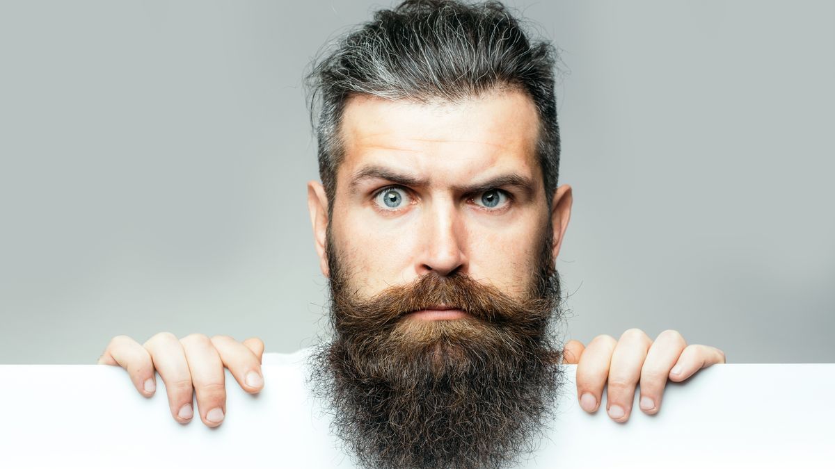 handsome bearded man with long lush beard and moustache on surprised face with white paper sheet in studio on grey background, copy space