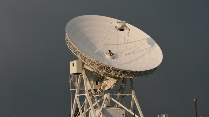 Duża biała antena satelitarna obracająca się na tle ciemnoszarego nieba, otoczona zielonymi polami.