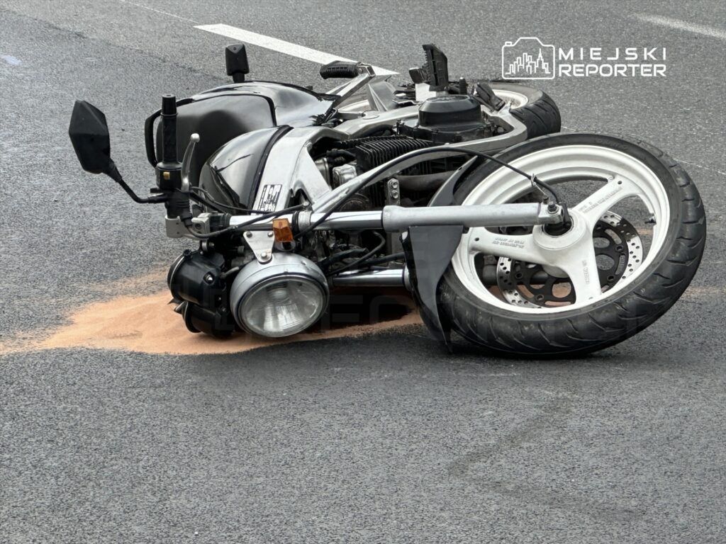 Motocykl leżący na boku na drodze, z plamą piasku obok, w miejskim otoczeniu.
