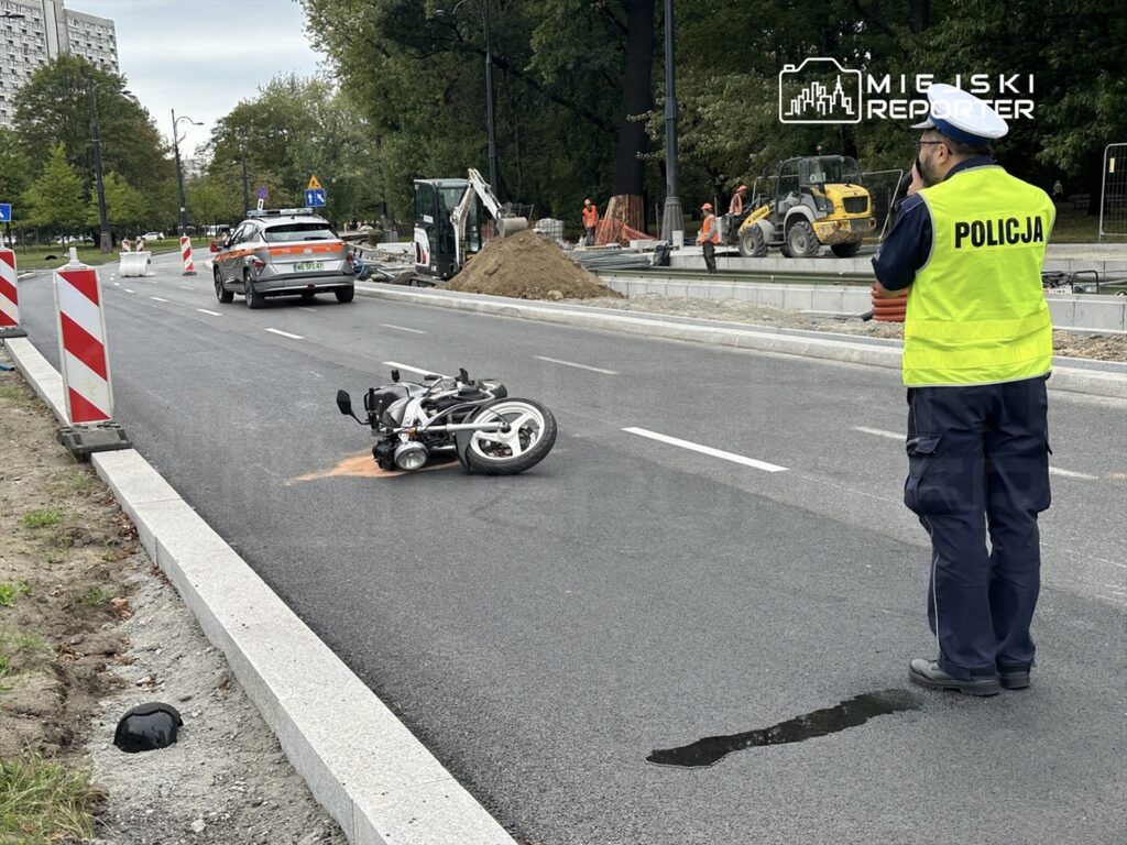 Motocykl leży na drodze, a policjant w kamizelce odblaskowej obserwuje miejsce zdarzenia w trakcie robót drogowych.