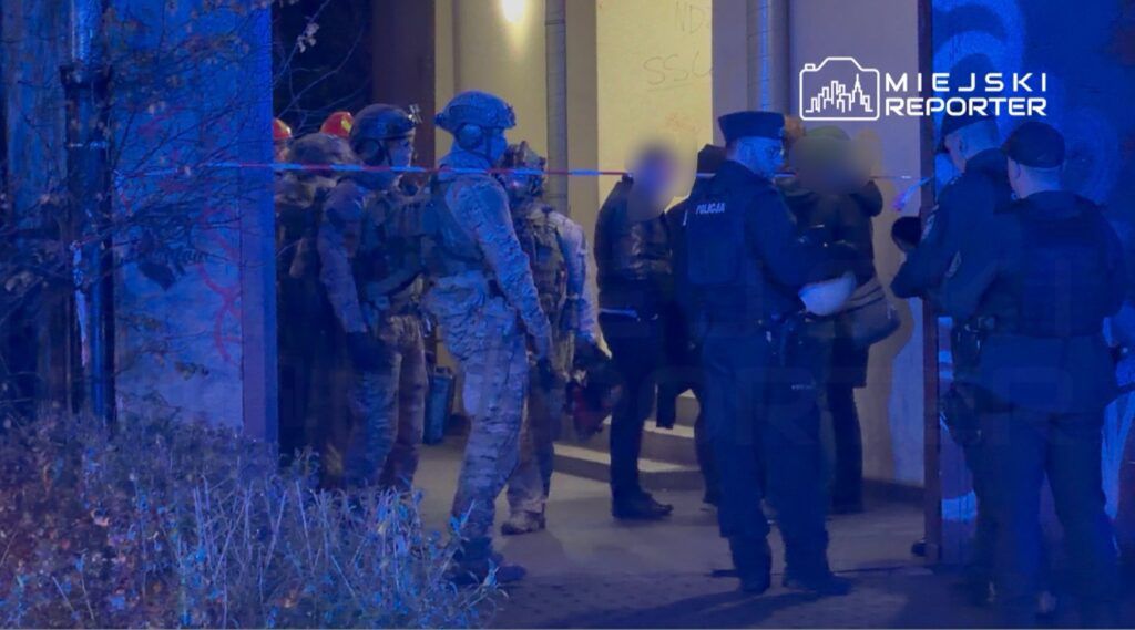 Grupa uzbrojonych policjantów i żołnierzy prowadzi działania w ciemnym otoczeniu budynku, zabezpieczając miejsce zdarzenia.