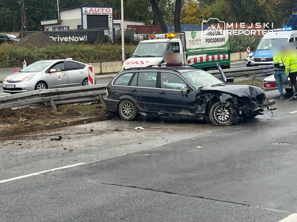 Czarny samochód osobowy z uszkodzeniami stoi przy barierce na drodze, obok interweniujący policjanci i służby.
