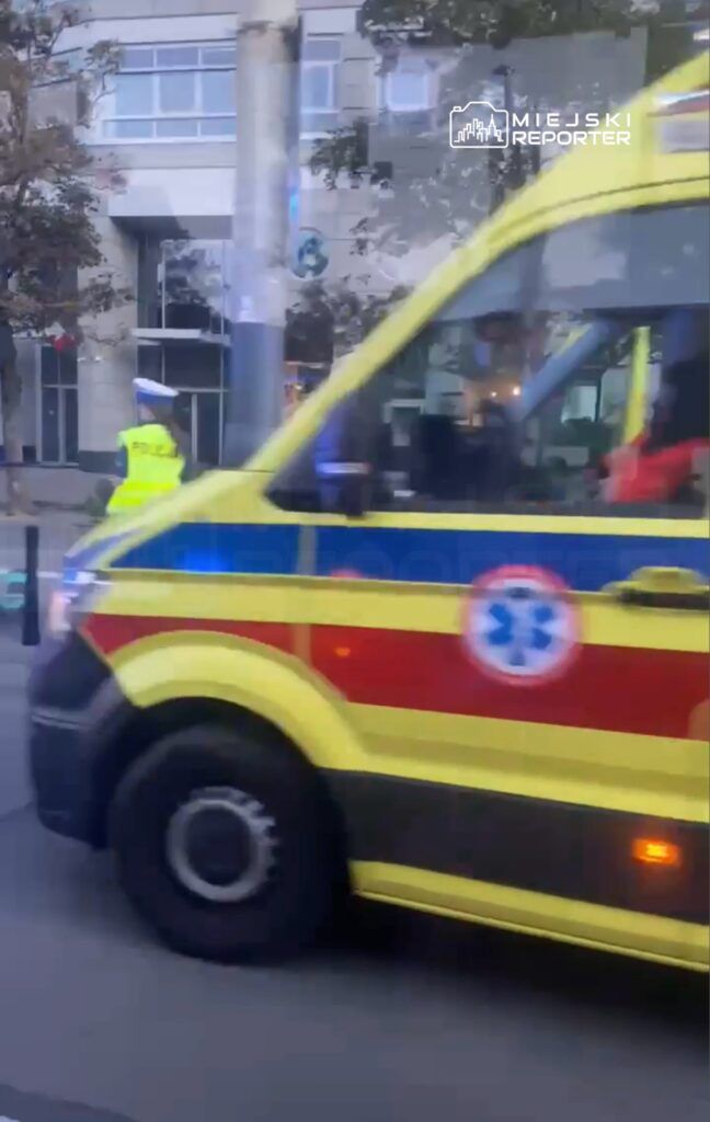 Karetka pogotowia przejeżdża obok policjanta kierującego ruchem na ulicy w miejskim otoczeniu.