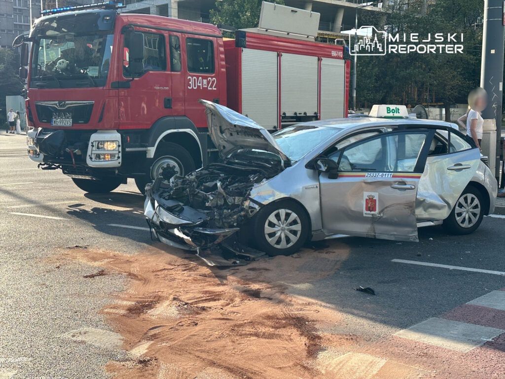 W zderzeniu drogowym uszkodzony samochód osobowy z otwartymi drzwiami stoi na jezdni, obok widać wóz strażacki.