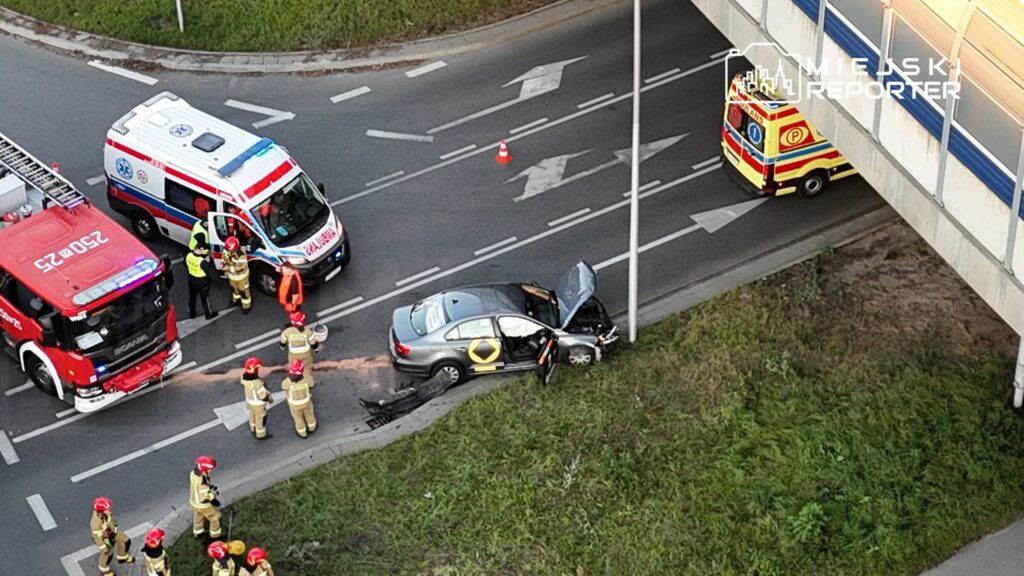 Na skrzyżowaniu stoi uszkodzony samochód osobowy, obok pracują strażacy i ratownicy medyczni.