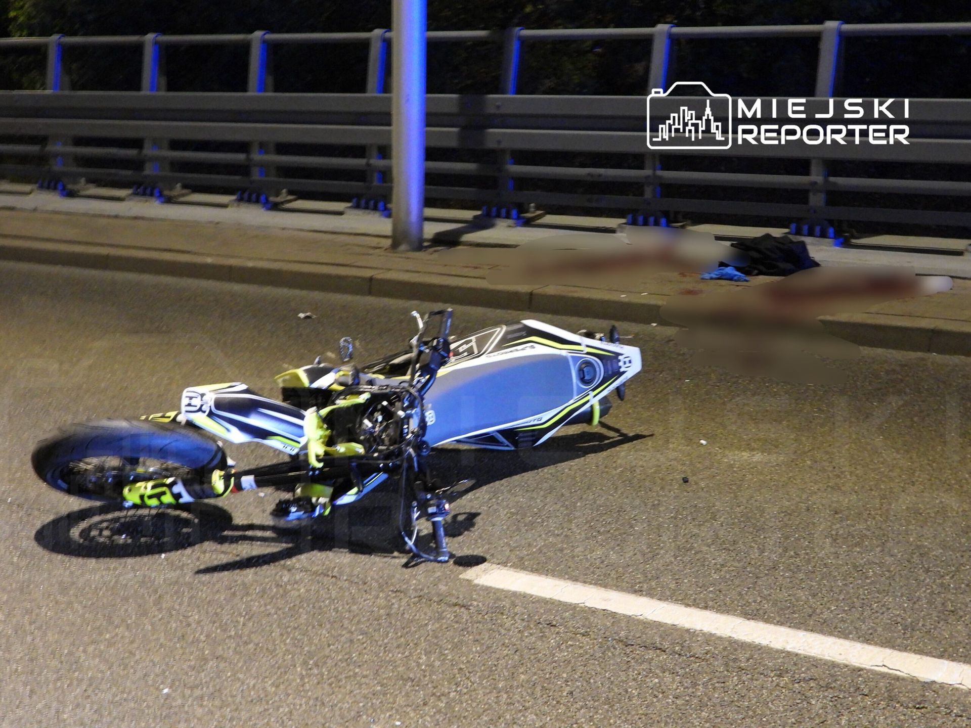 Motocykl leżący na jezdni, obok widoczne plamy krwi oraz ciuchy. Nocne oświetlenie ulicy.