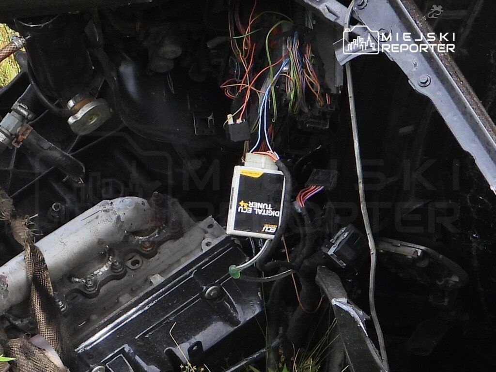 Uszkodzony silnik samochodowy z widocznymi przewodami elektrycznymi oraz modułem sterującym w otoczeniu trawy.