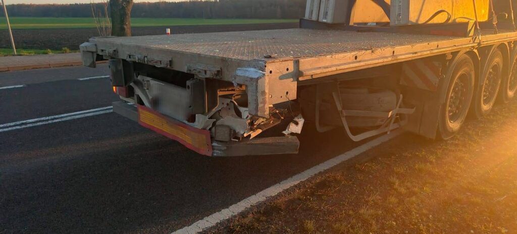 Uszkodzona tylna część naczepy ciężarówki zaparkowanej na poboczu drogi, w tle widoczne pola i drzewa.