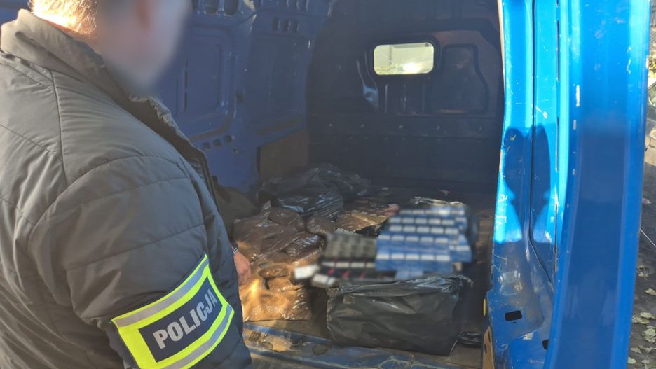 Policjant w mundurze sprawdzający zawartość niebieskiego furgonu, w którym znajdują się torby z nieznanym ładunkiem.