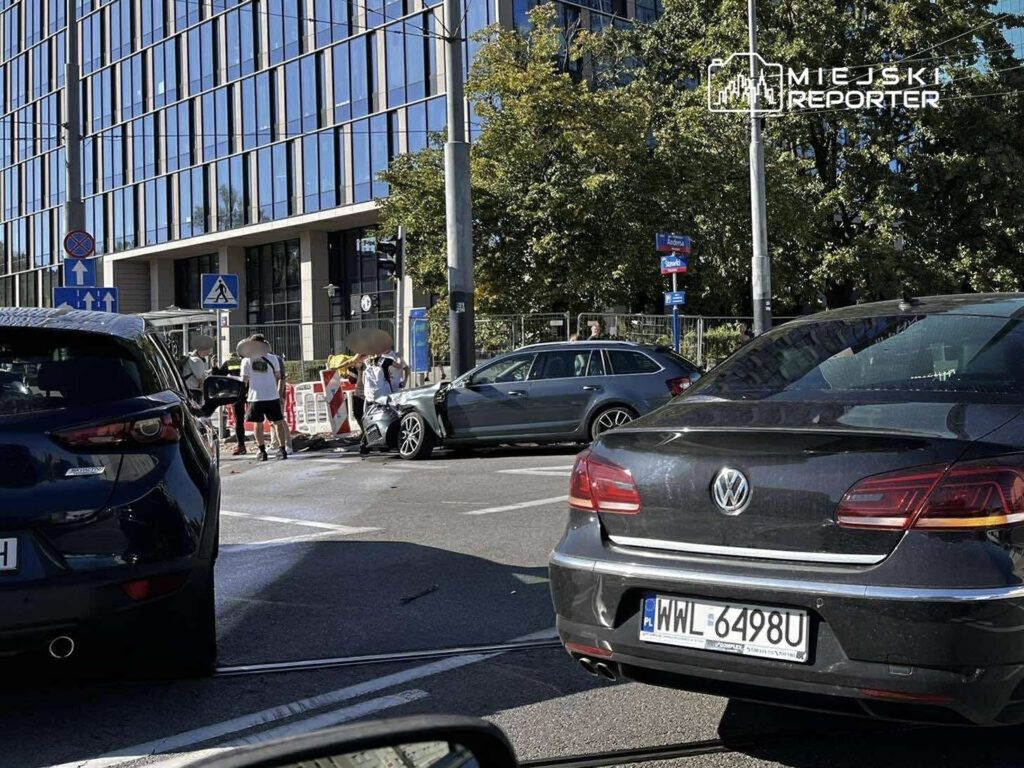 Grupa osób w białych koszulkach ocenia uszkodzenia dwóch samochodów po kolizji na ruchliwej ulicy obok nowoczesnego biurowca.