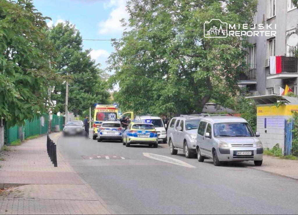 Na ulicy zaparkowane są pojazdy, w tym karetka pogotowia i policyjny radiowóz, a funkcjonariusze interweniują.