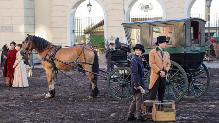 Mężczyzna w kapeluszu i płaszczu stoi obok konia zaprzęgowego, a obok niego dwie osoby w strojach historycznych.