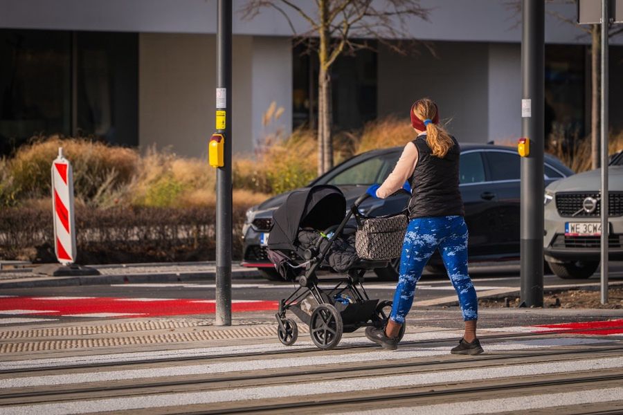 Kobieta w legginsach i kamizelce prowadzi wózek dziecięcy przez przejście dla pieszych.