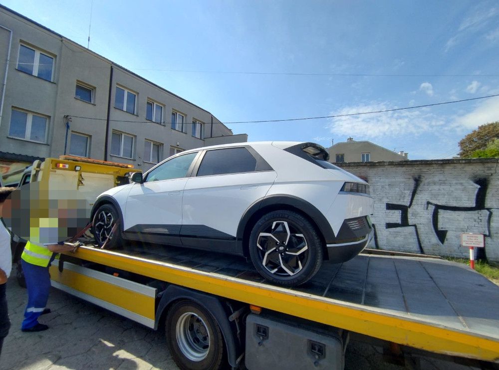 Biały samochód elektryczny załadunkowy na platformie lawety w miejskim otoczeniu z graffiti w tle.