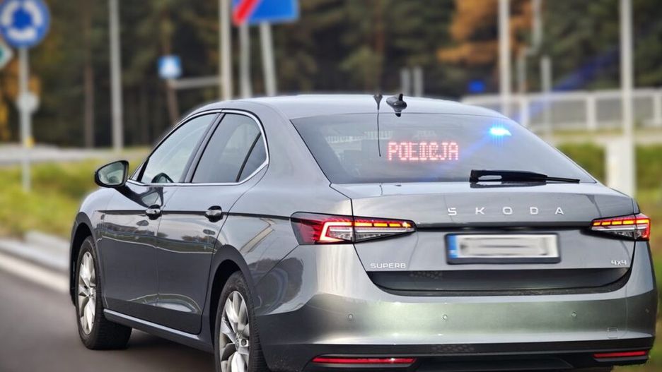 Radiowóz policyjny marki Skoda z sygnalizacją świetlną zatrzymany na poboczu drogi, z widocznym napisem "POLICJA" na tylnej szybie.